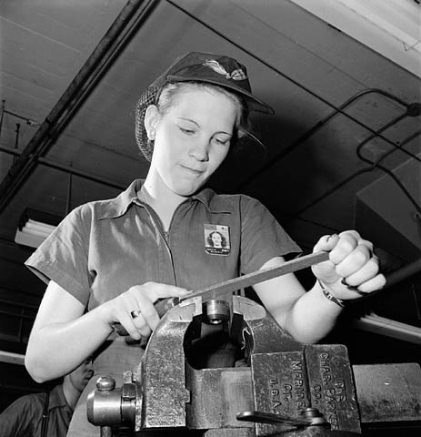 Lady factory worker files a machine element while piped music plays on loudspeakers.